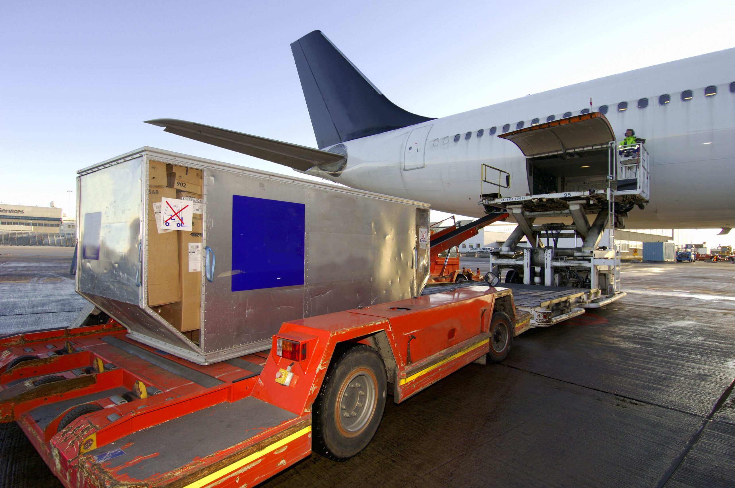 A large metal crate on a red baggage cart is about to be loaded into an airplane's cargo hold.