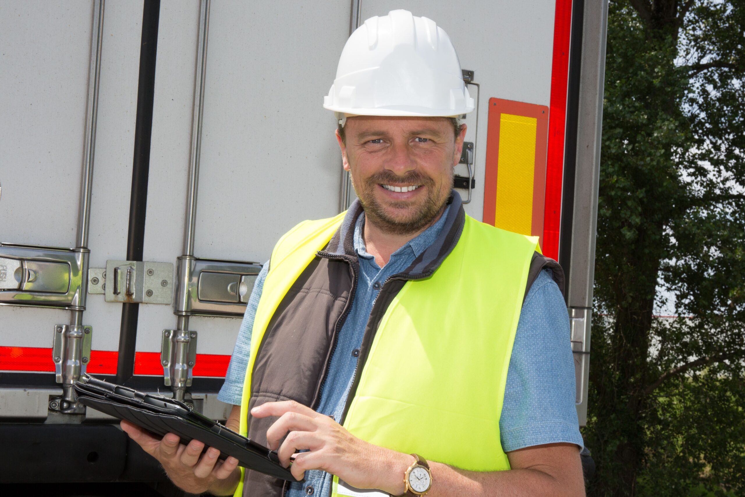 A man in a hard hat and reflective vest smiles while using a tablet in front of a truck, suggesting a worksite setting with a positive tone.