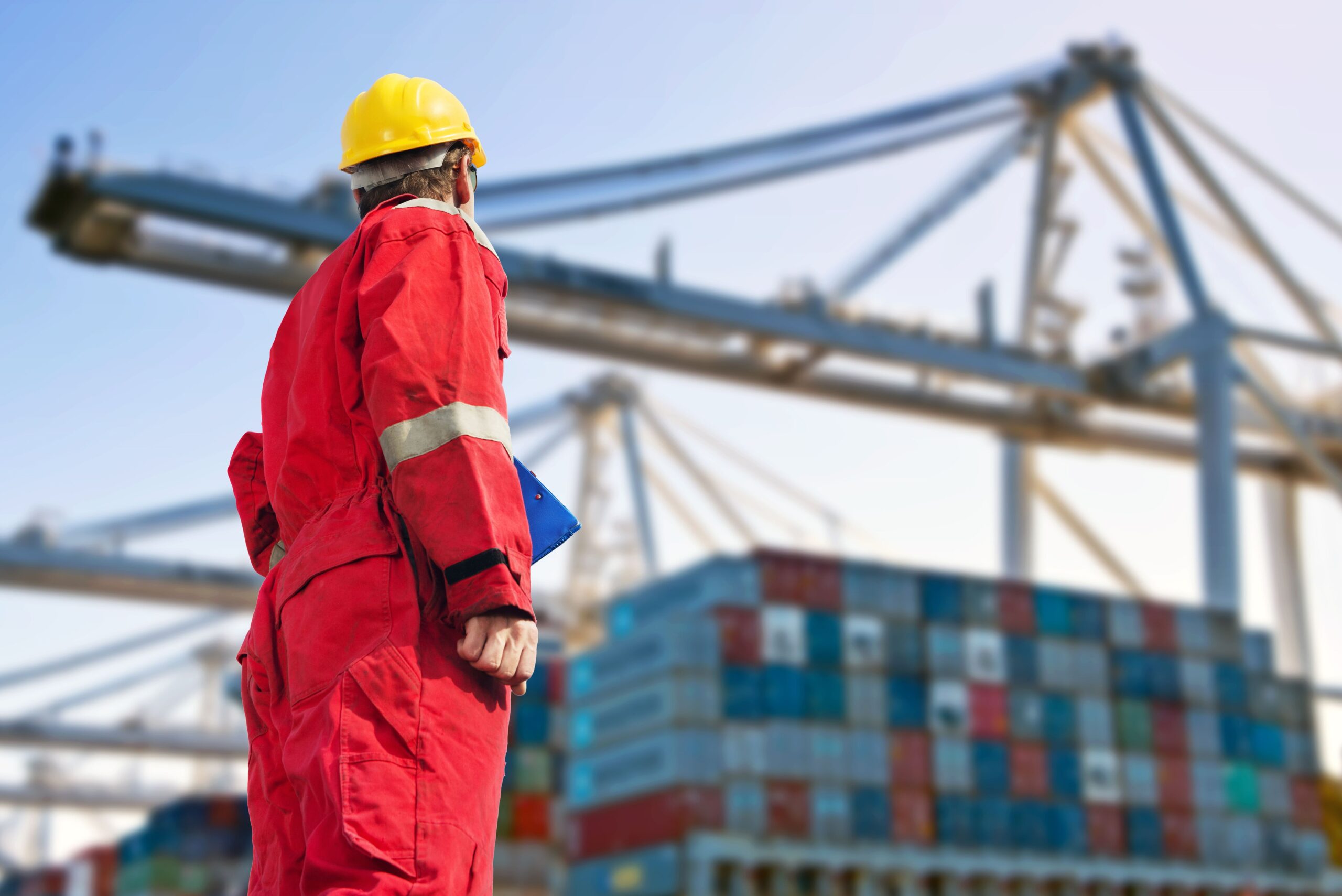 A worker in a red jumpsuit and yellow hard hat stands in a shipping yard, holding a clipboard, with stacks of colorful containers and cranes behind.