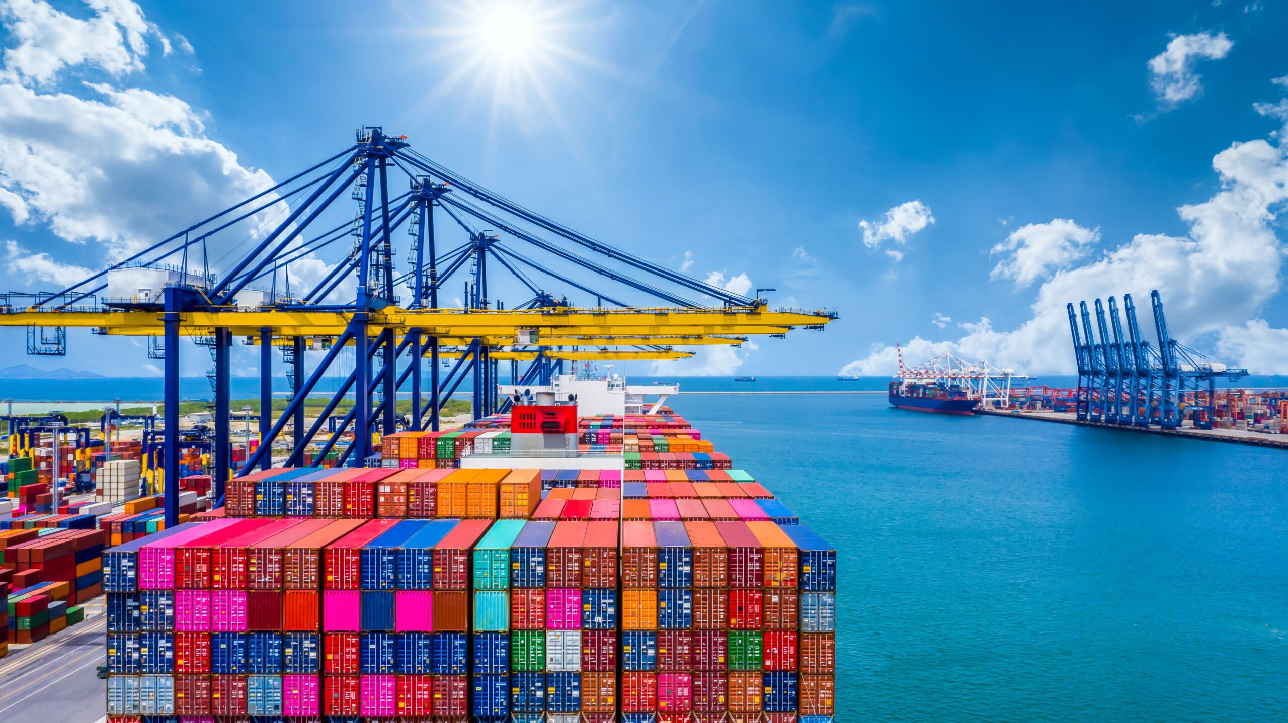 A vibrant, busy seaport under a bright blue sky shows rows of colorful shipping containers on a large cargo ship, with cranes operating nearby.