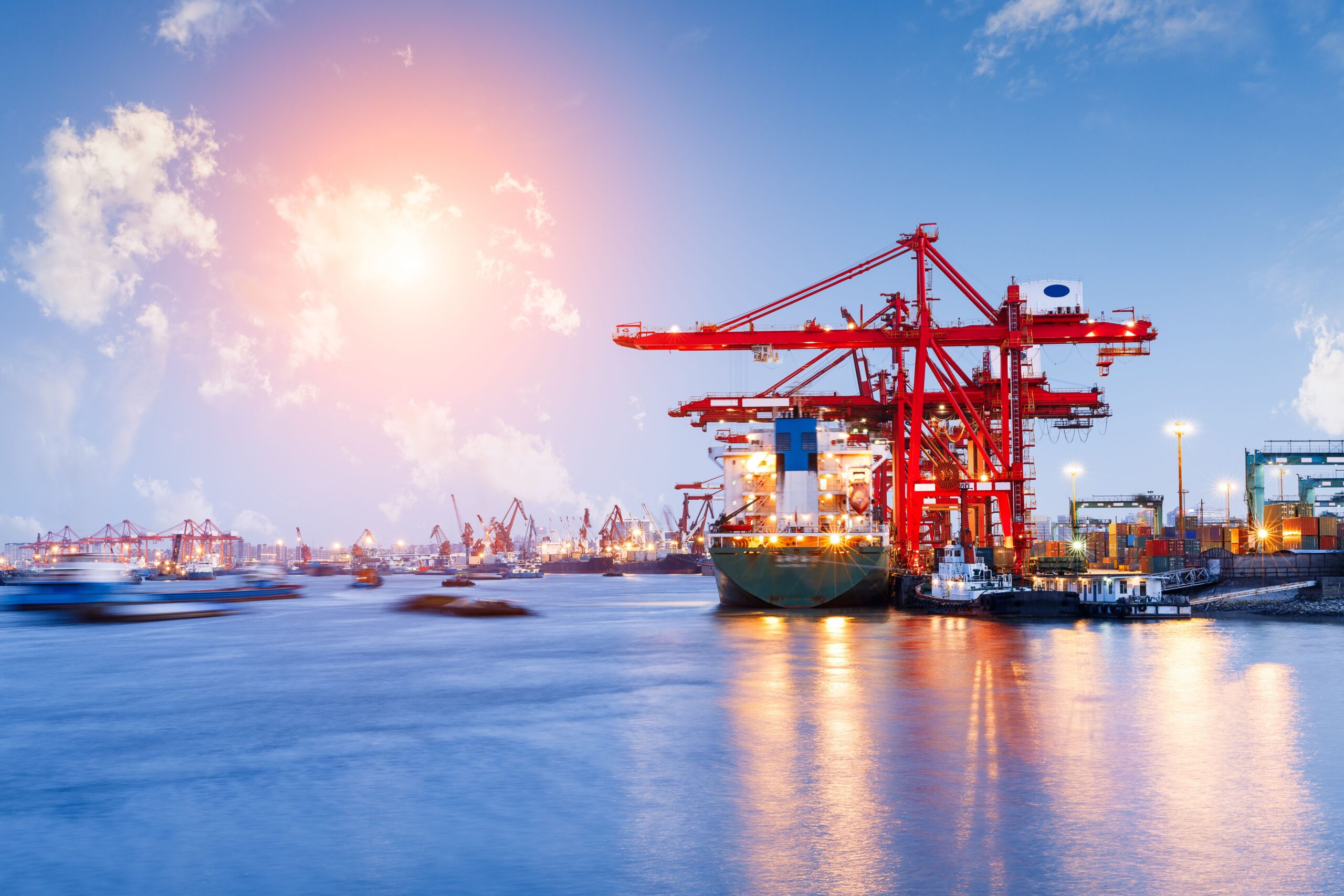 A bustling port with red cranes unloads a cargo ship under a vibrant sky at sunset.